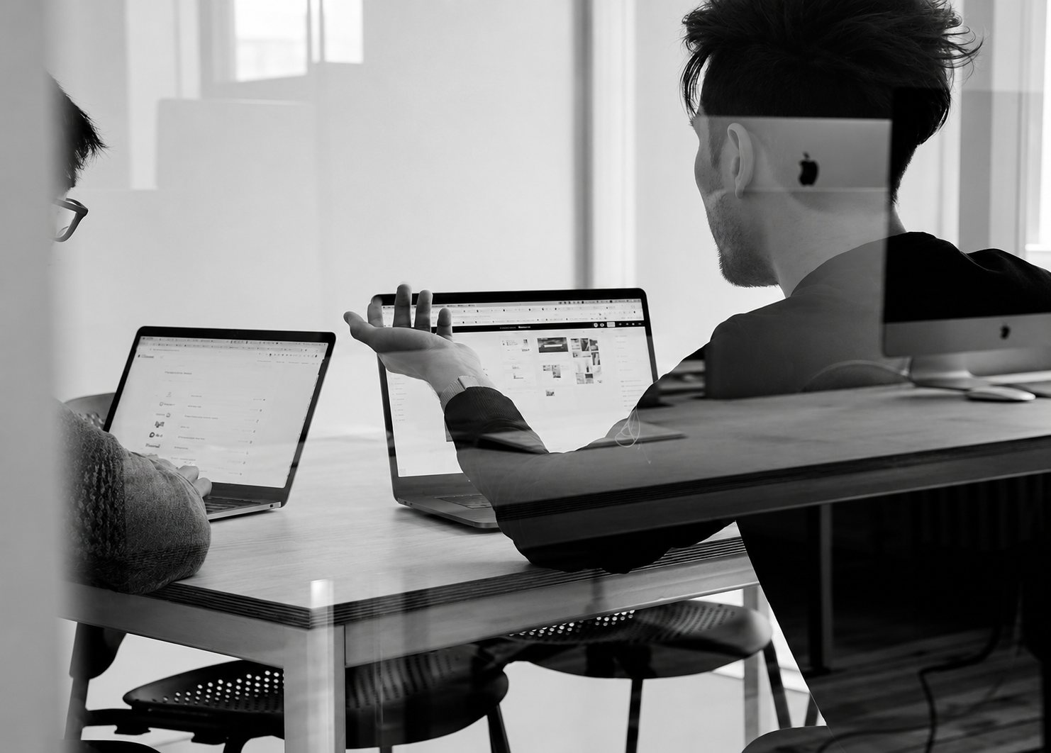 Photo en noir et blanc, prise à travers une vitre, de deux personnes assises à une table de travail devant des ordinateurs portables, l'une faisant un geste de la main.