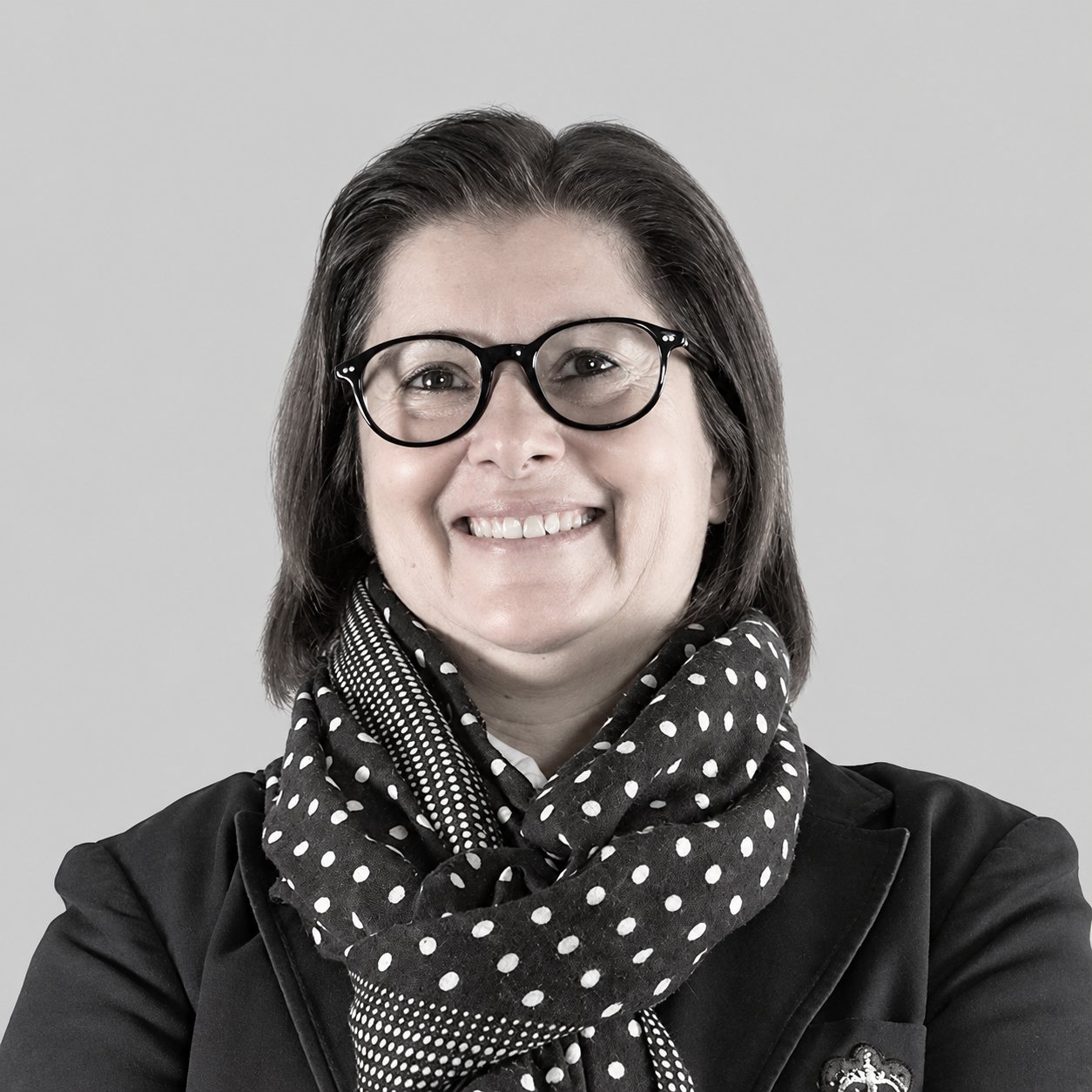 Portrait en noir et blanc d'une femme souriante portant des lunettes rondes, un foulard à pois et une veste sombre devant un fond gris uni.