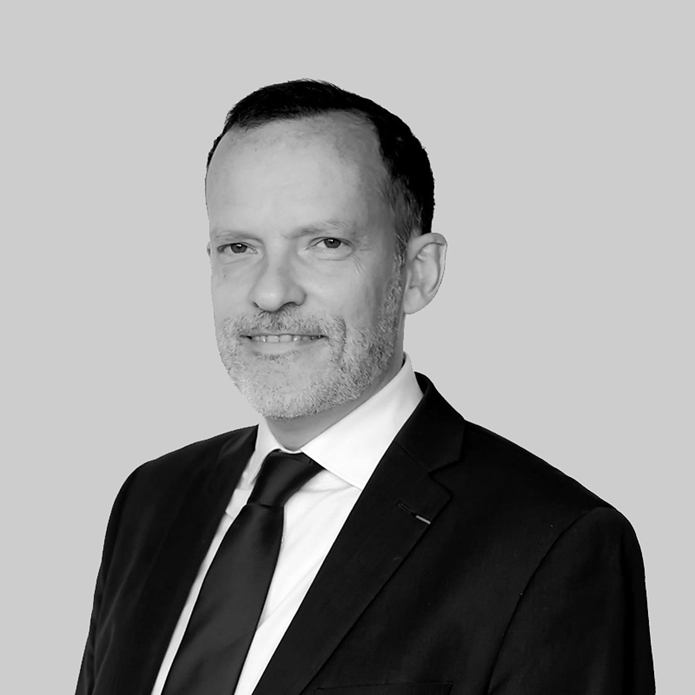 Portrait en noir et blanc d'un homme souriant à la barbe grisonnante, vêtu d'un costume sombre, d'une chemise blanche et d'une cravate, devant un fond gris uni.