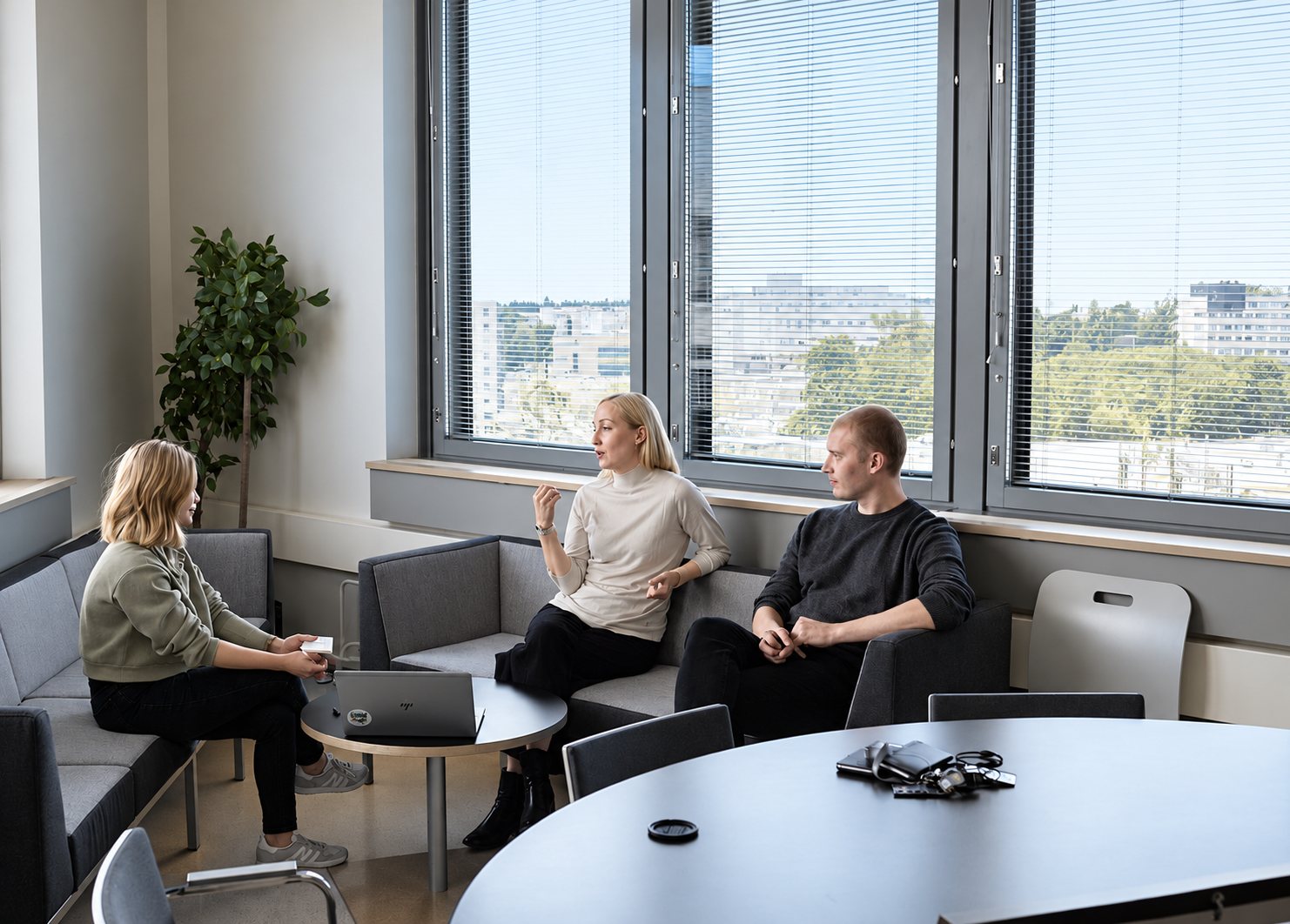 Photo en noir et blanc de trois personnes assises dans un coin salon de bureau devant de grandes fenêtres, en pleine discussion autour d'un ordinateur portable.